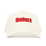 The Nebraska Sideline Hat by No Rivals is a white snapback with "Huskers" embroidered in red on the front. This structured cotton cap is shown against a white background.