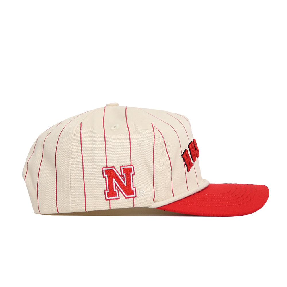 The No Rivals Nebraska Walk Off Hat is a cream cotton snapback with vertical red pinstripes, a red brim, a side N logo, and partially visible red front lettering. Officially licensed, it blends style and comfort.