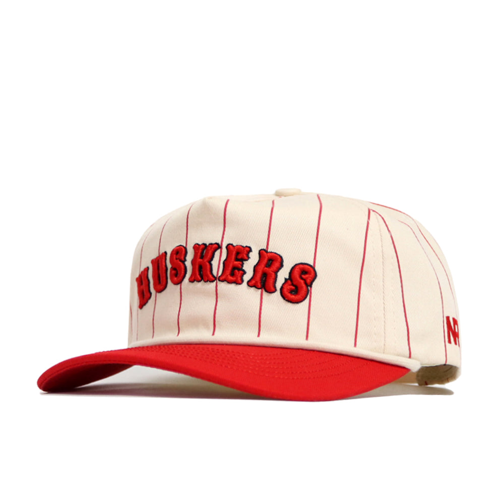 The Nebraska Walk Off Hat by No Rivals features cream cotton with red pinstripes, a red brim, and "HUSKERS" embroidered in red with a black outline. This adjustable cap offers comfort and classic style.