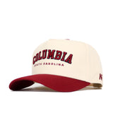 The Columbia Hometown Hat by No Rivals is a cream baseball cap with a red brim and structured fit. "COLUMBIA" in bold red and black, with "SOUTH CAROLINA" below in smaller red text, is featured on the front. Made from soft cotton fabric.