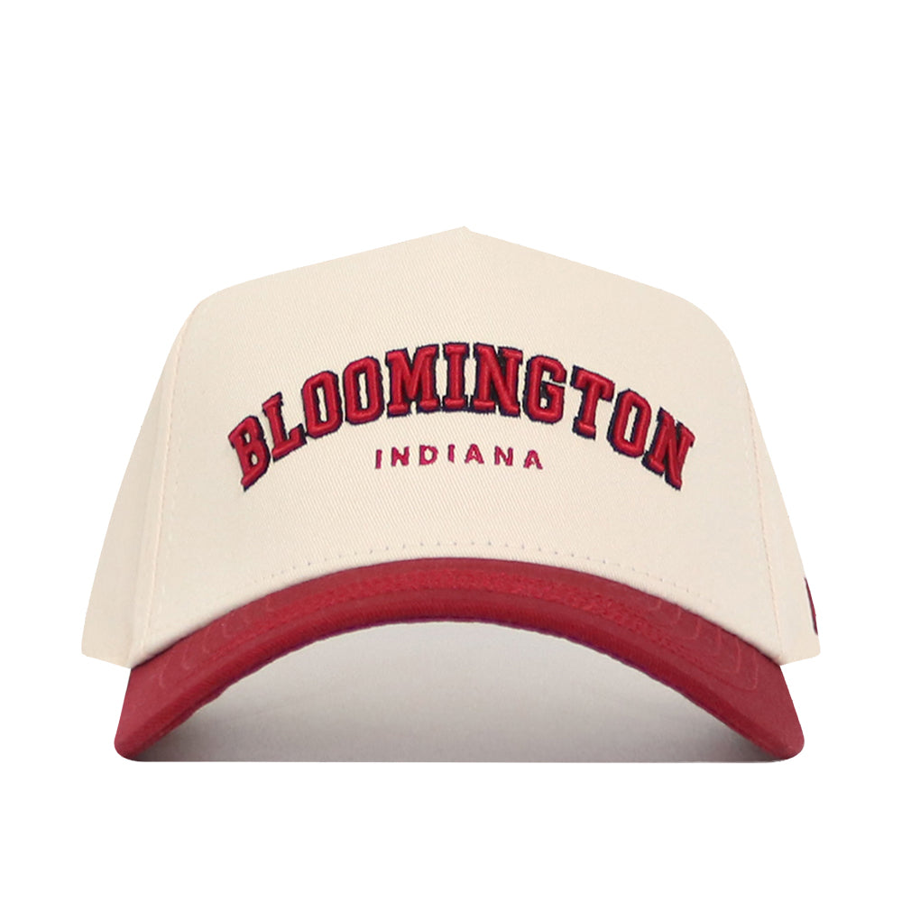 The No Rivals Bloomington Hometown Hat is a cream cotton snapback cap with a red brim, featuring "BLOOMINGTON" in large red and black letters above "INDIANA" in smaller red letters on the front.