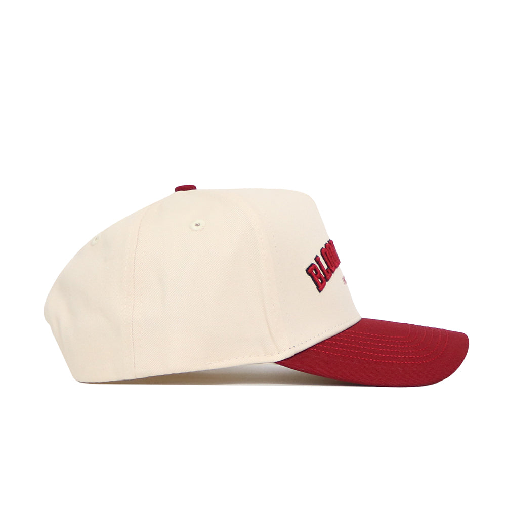 The No Rivals Bloomington Hometown Hat is a cream cotton cap with a curved maroon brim, side view. It features ventilation eyelets, adjustable fit, and partially visible maroon embroidery on the front.