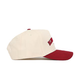 The No Rivals Bloomington Hometown Hat is a cream cotton cap with a curved maroon brim, side view. It features ventilation eyelets, adjustable fit, and partially visible maroon embroidery on the front.