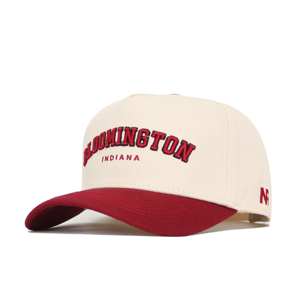The No Rivals Bloomington Hometown Hat is a cream cotton adjustable cap with a red brim. "Bloomington" in large red letters outlined in black and "Indiana" below are embroidered on the front, with a small red logo on the side.