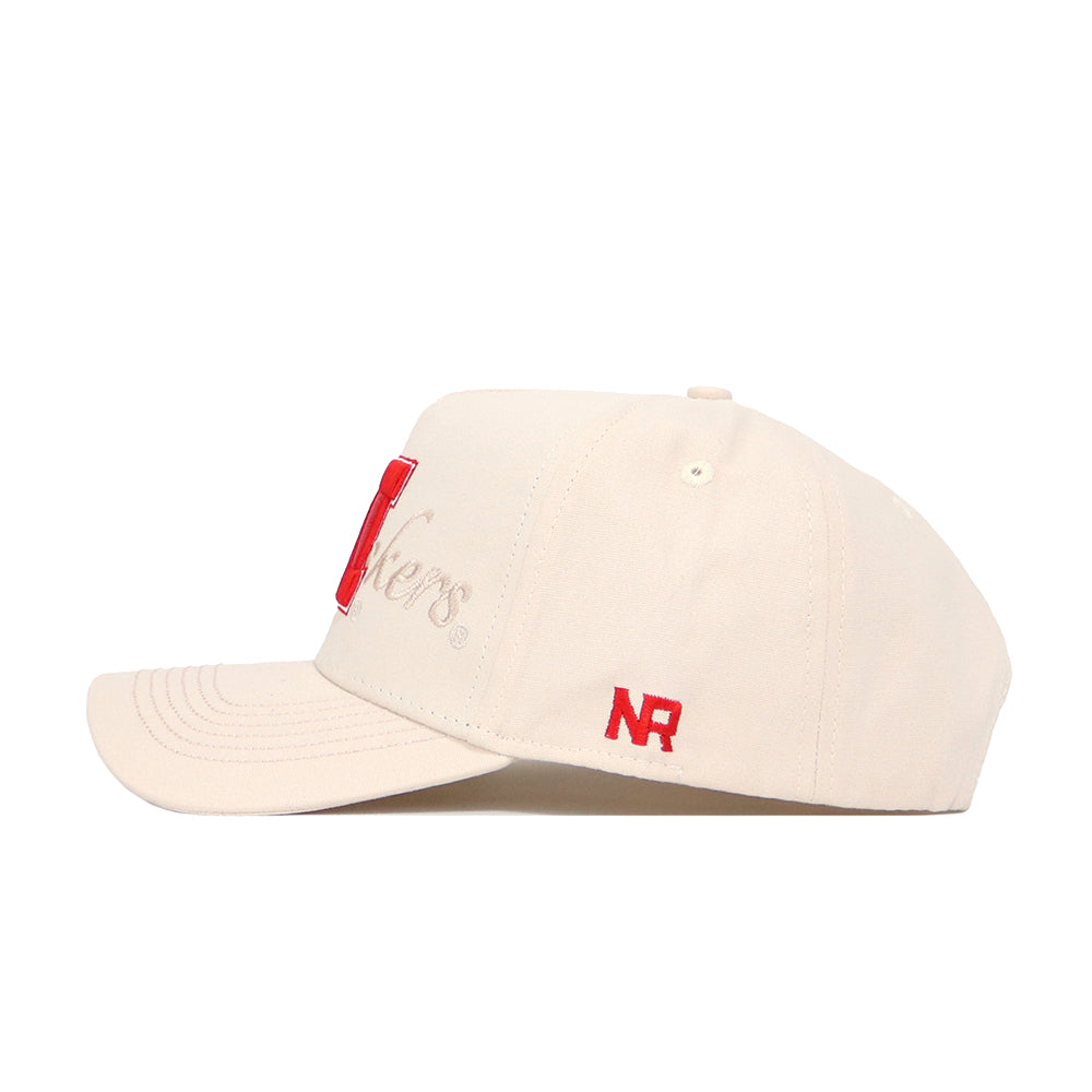 The Nebraska Stacked Hat by No Rivals is a beige snapback made from premium cotton, with red NR embroidery on the side and "Huskers" partly on the front. Its curved brim and ventilation eyelets provide a comfortable, structured fit.