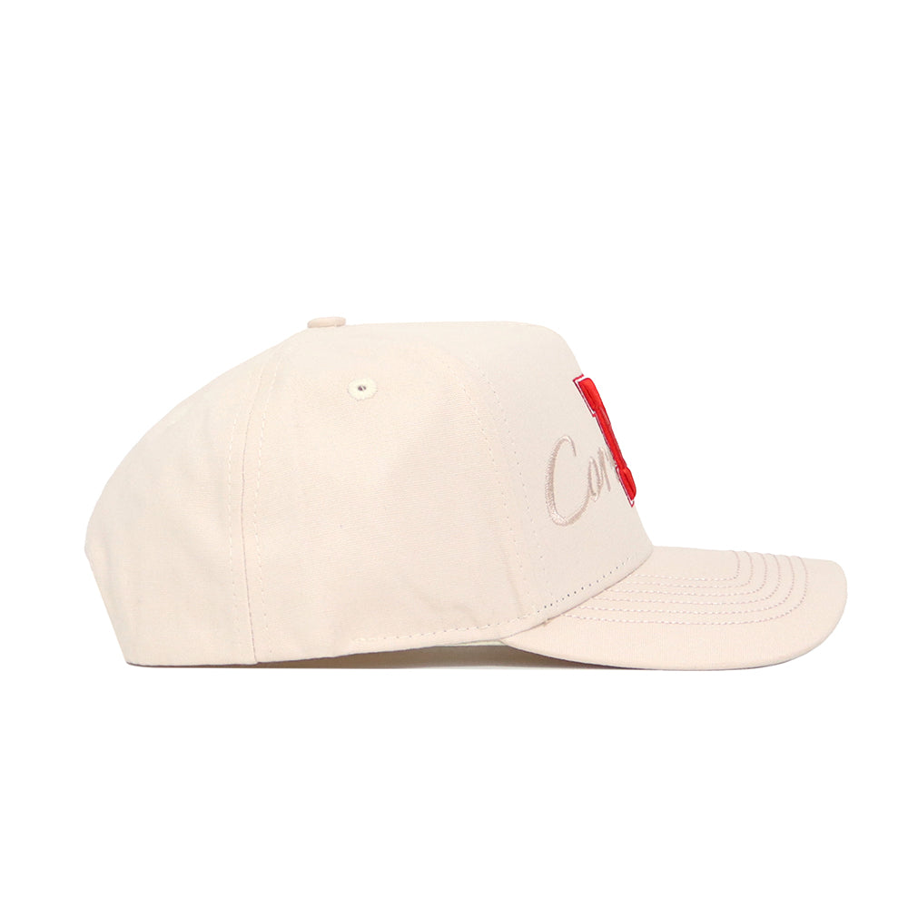 The Nebraska Stacked Hat by No Rivals is a beige cotton baseball cap, viewed from the side, with subtle embroidered text and a bold red rectangular patch on the front panel.
