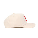 The Nebraska Stacked Hat by No Rivals is a beige cotton baseball cap, viewed from the side, with subtle embroidered text and a bold red rectangular patch on the front panel.