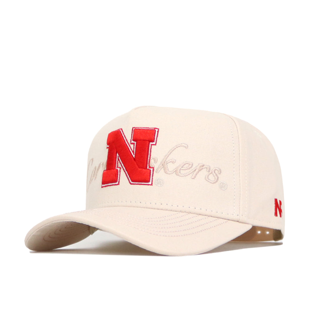 The No Rivals Nebraska Stacked Hat is a cream-colored, structured-fit cotton hat featuring a large red N on the front, “Cornhuskers” embroidered in script, and a small red N on the side.