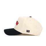 The Athens Hometown Hat by No Rivals is a cream-colored snapback baseball cap with a black curved brim, made from quality cotton. It features NR initials embroidered on the side and red letters partially visible on the front.