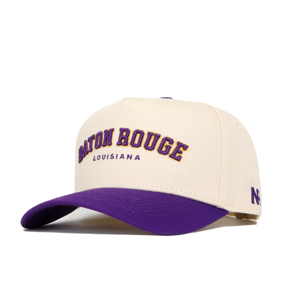 The No Rivals Baton Rouge Hometown Hat is a cream cotton baseball cap with a structured fit, purple brim, and "Baton Rouge" embroidered in large purple and gold letters above "Louisiana" in small purple text on the front.