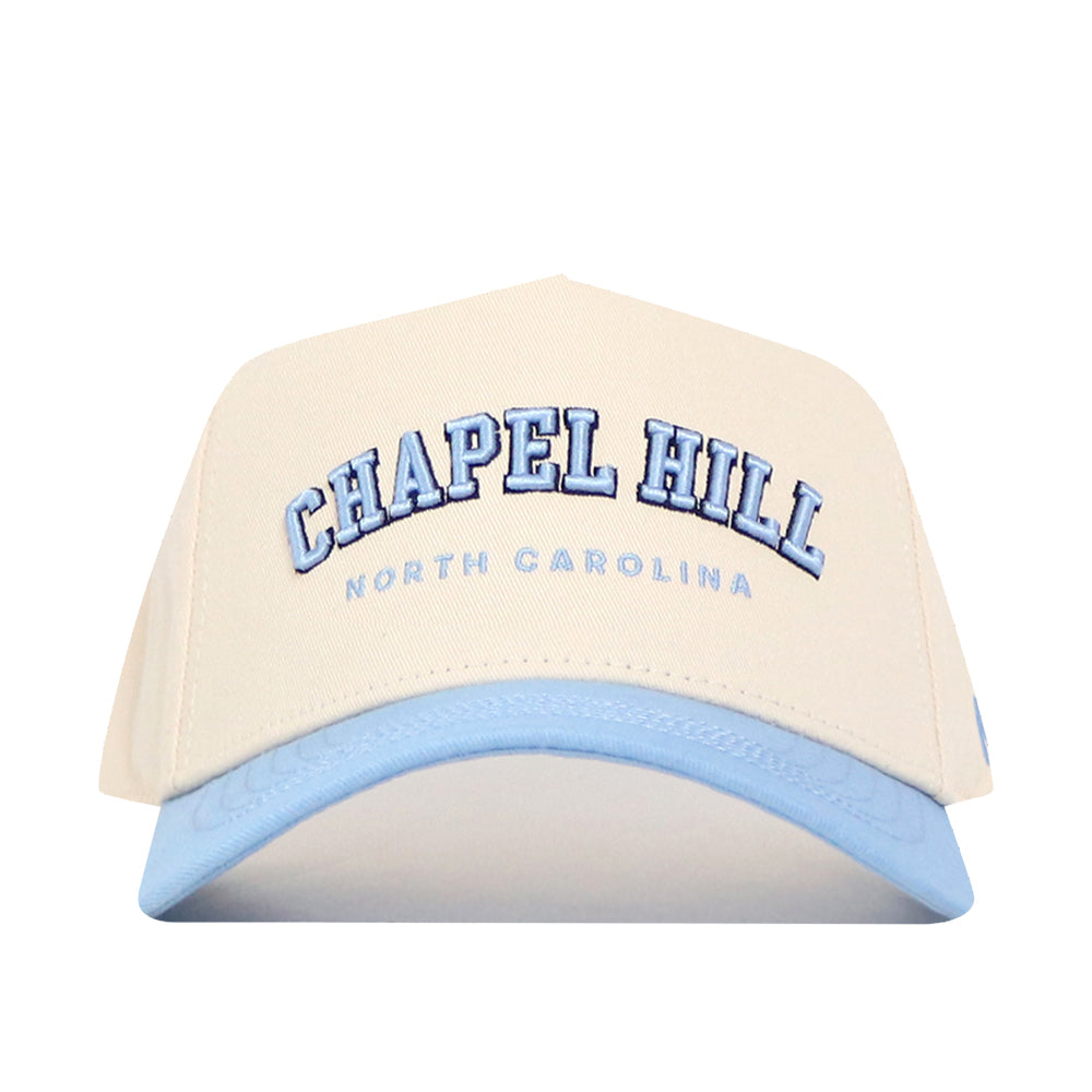 The No Rivals Chapel Hill Hometown Hat is a beige cotton cap with a light blue brim, featuring "CHAPEL HILL" in large blue letters and "NORTH CAROLINA" below.
