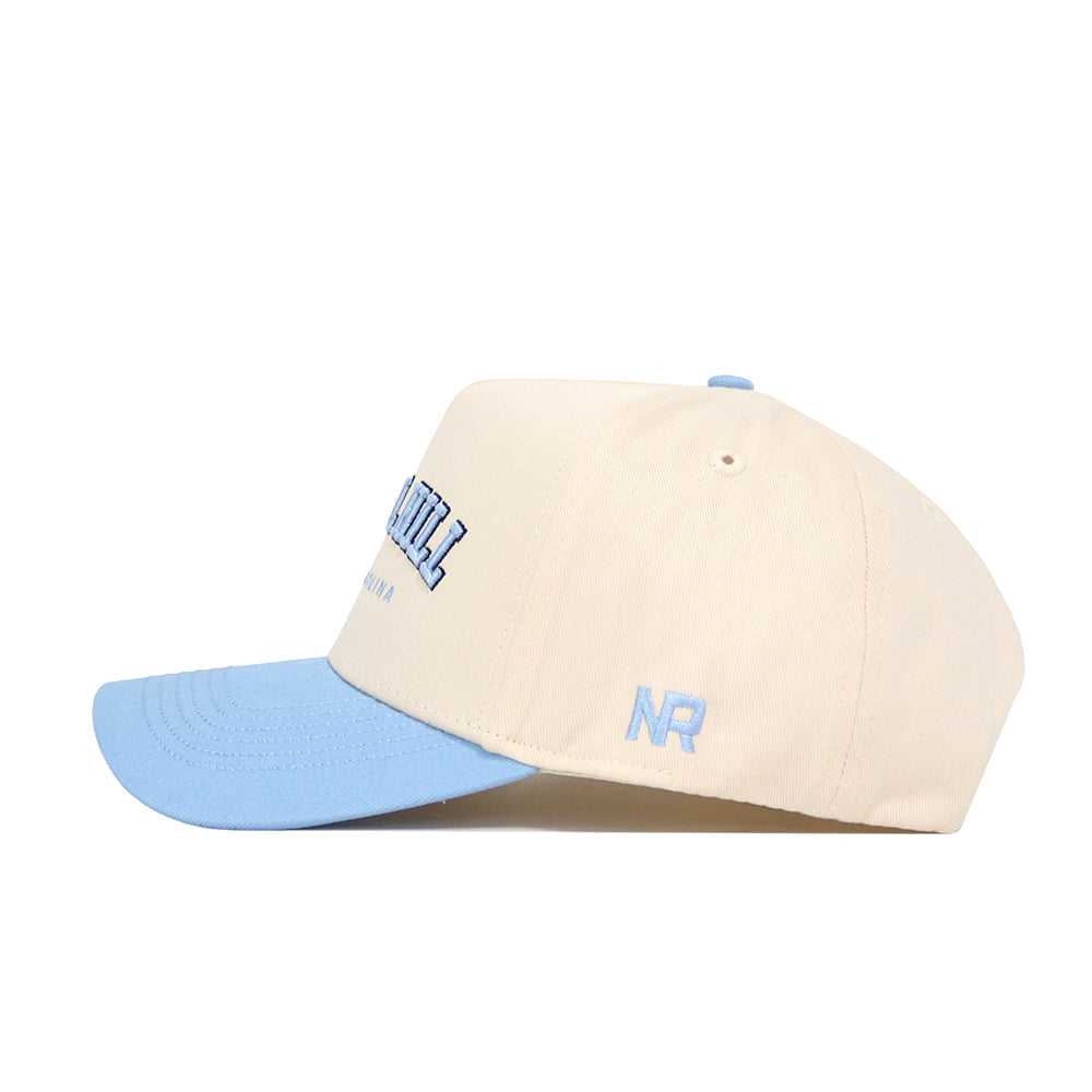 The No Rivals Chapel Hill Hometown Hat is a cream snapback with a light blue brim, blue embroidered front text, and NR initials on the side. Made from cotton for comfort and a structured fit.