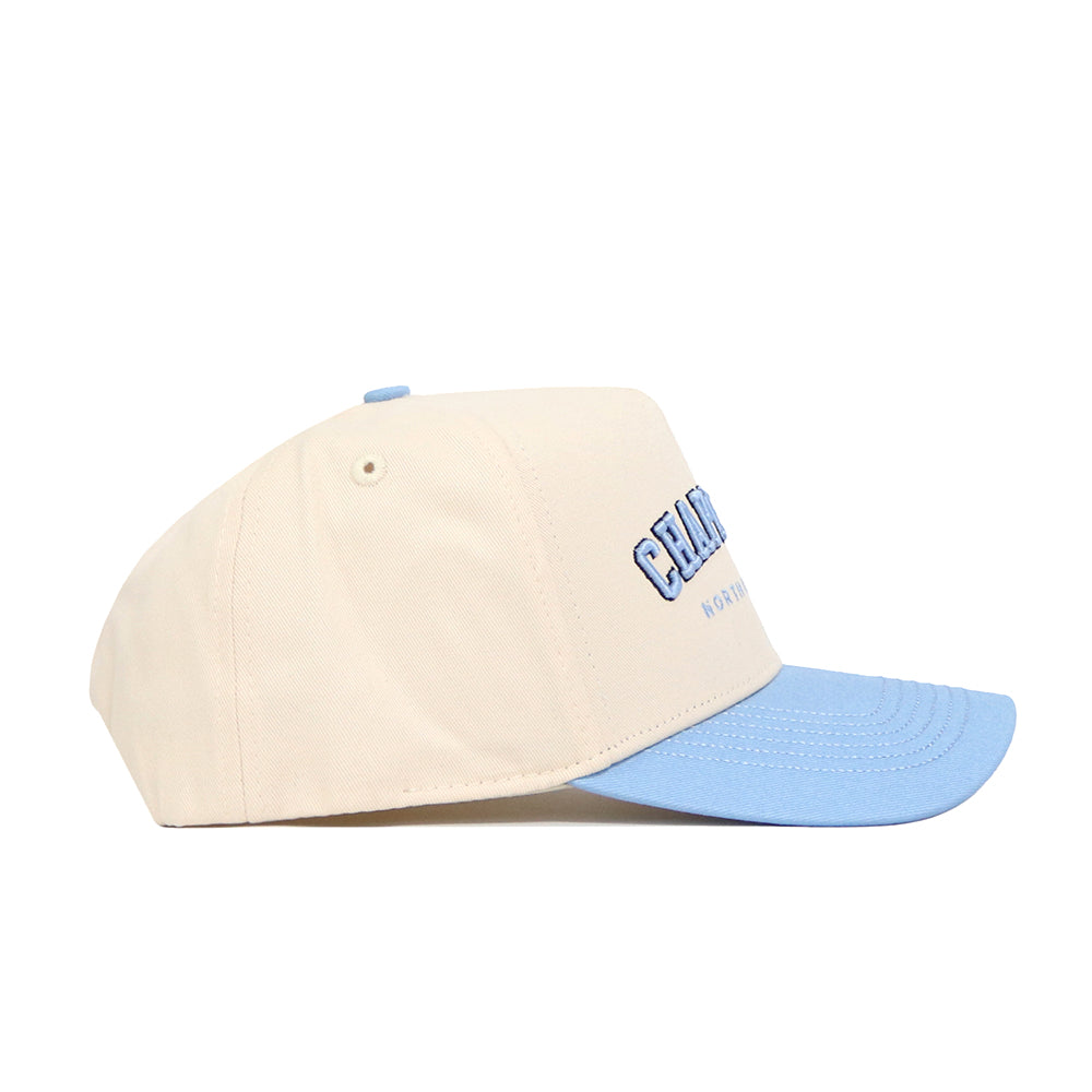 The No Rivals Chapel Hill Hometown Hat is a beige snapback with a structured fit, light blue brim, and blue embroidered front text, shown from the side.