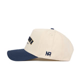 The No Rivals Morgan Town Hometown Hat is a structured cream cap with a navy brim, front embroidery, NR initials on the side, high-quality cotton construction, and a classic snapback closure.