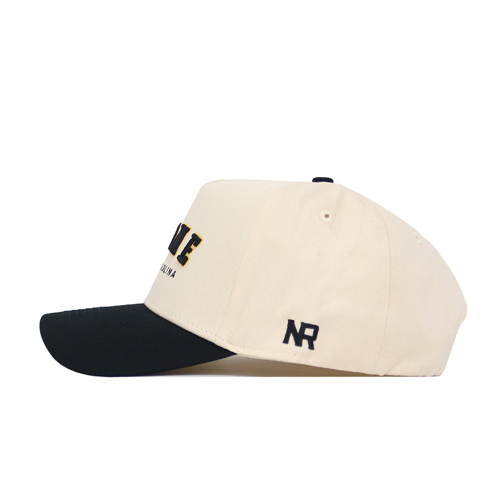 The Boone Hometown Hat by No Rivals is a cream-colored baseball cap with a black brim, made of high-quality cotton, featuring black embroidered NR letters on the side and partially visible text on the front.