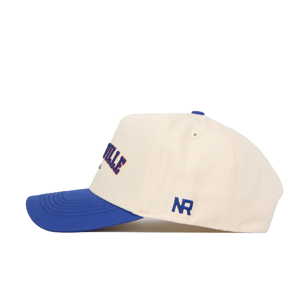 The Gainesville Hometown Hat by No Rivals is a cream baseball cap with a blue brim and top button, embroidered blue NR initials on the side, and red-blue lettering on the front. Made from cotton with a structured fit.