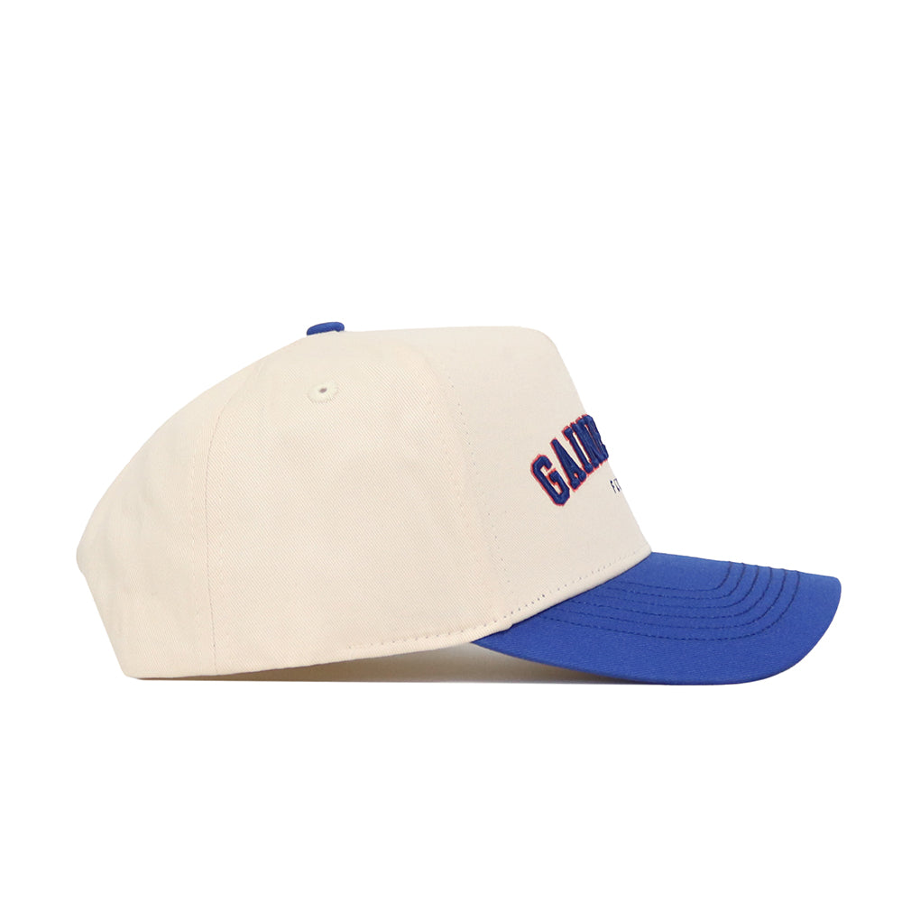 The No Rivals Gainesville Hometown Hat is a cream cotton baseball cap with a blue brim, shown from the side. It has a structured fit and features embroidered blue and red text on the front panel.