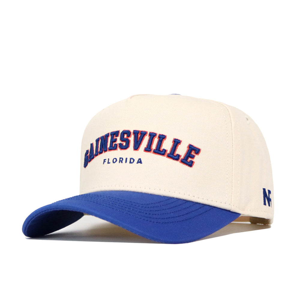 The Gainesville Hometown Hat by No Rivals is a beige cotton baseball cap with a blue brim, bold blue and red "GAINESVILLE FLORIDA" on the front, structured fit, and a small blue NF logo on the side.