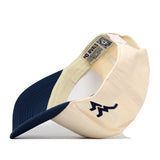 A cream and navy blue WVU Mountaineers Mascot Patch Hat by No Rivals is displayed from the side and inside, featuring a zigzag logo on the front and an interior label that reads NO RIVALS. This officially licensed cap includes a convenient snap back closure for an adjustable fit.