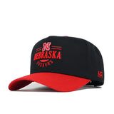The No Rivals Nebraska Tailgate Hat is a black snapback with a curved red brim, featuring red and white embroidered N Nebraska Huskers on the front and a small red N on the side. Made from high quality cotton with a structured fit.