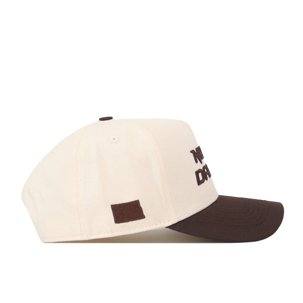 A side view of the North Dakota Espresso Hat by No Rivals shows a cream-colored baseball cap with a dark brown bill, a small brown rectangle on the side, and partially visible dark brown embroidered logo text on the front.