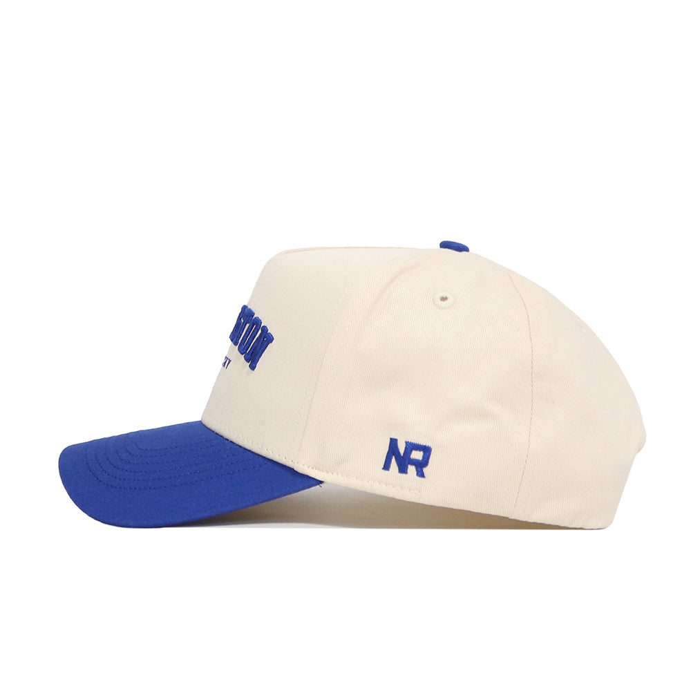 The No Rivals Lexington Hometown Hat is cream-colored cotton with a blue brim, blue stitched NR on the side, matching stitching, ventilation eyelets, and an adjustable snap-back for a structured, customizable fit.