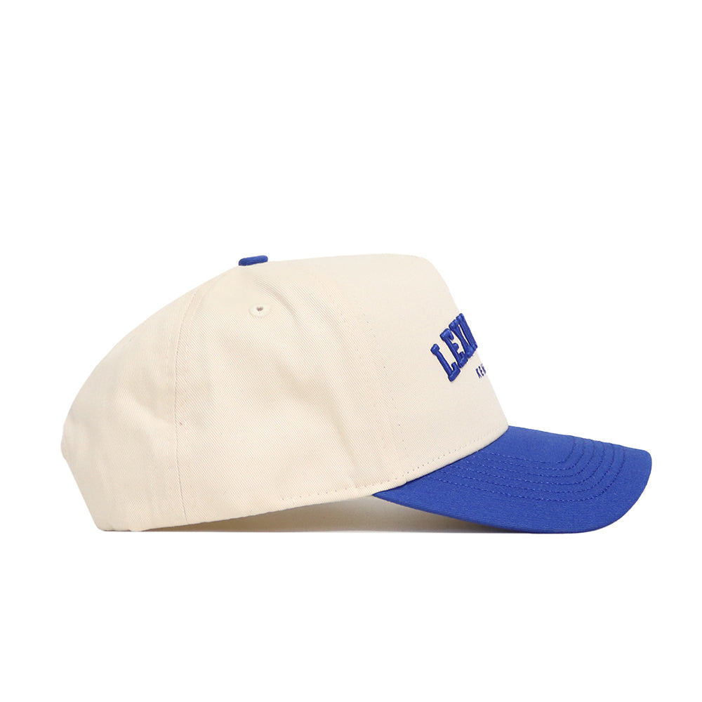 The Lexington Hometown Hat by No Rivals is a cream, high-quality cotton hat with a blue brim and embroidered front letters. This structured fit features a snap back for adjustable comfort, shown here in side profile.