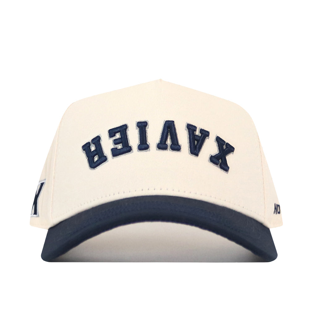 Show your support with the No Rivals Xavier Musketeers Upside Down Xavier Hat – Classic. This cream-colored snapback cap features a dark blue brim and the word XAVIER embroidered in navy blue letters upside down on the front, making it an officially licensed standout piece.