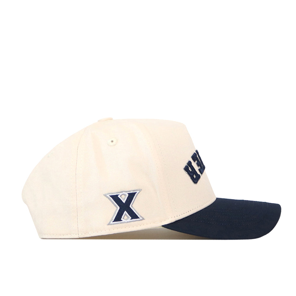 Introducing the Xavier Musketeers | Upside Down Xavier Hat - Classic by No Rivals: this officially licensed beige snapback features a navy blue brim, a distinctive blue and white “X” logo on the side, and partial black lettering on the front—blending team pride with classic style.