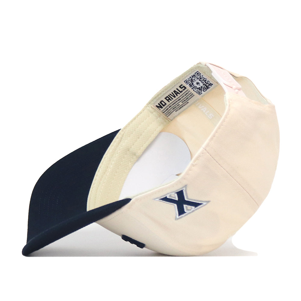 An officially licensed Xavier Musketeers | Upside Down Xavier Hat - Classic by No Rivals is shown upside down, featuring a cream and navy design with an X logo on the side and an interior label reading “NO RIVALS.” The inside of the brim and tag details are visible, showcasing its classic snapback styling.