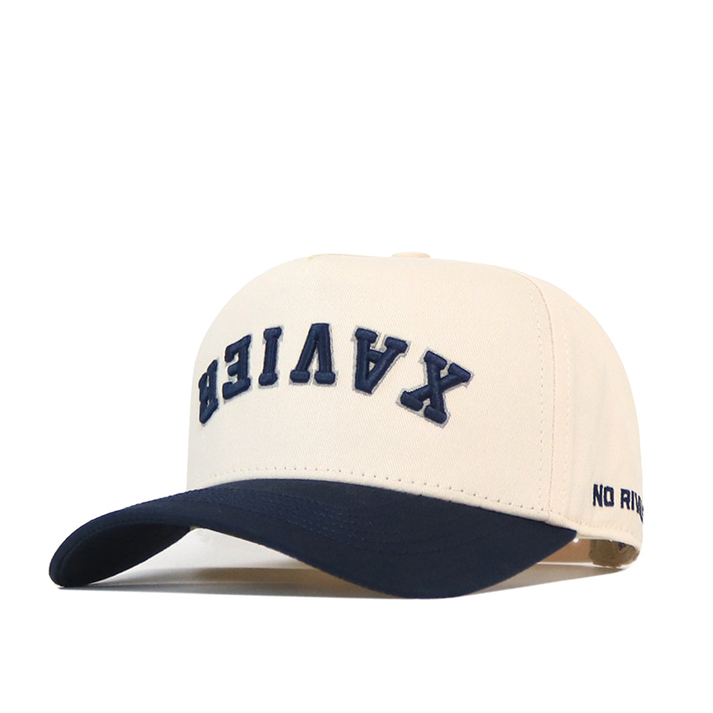 Show off your team pride with the officially licensed Xavier Musketeers | Upside Down Xavier Hat - Classic by No Rivals, a white and navy snapback featuring “XAVIER” in bold upside-down blue letters on the front and “NO RIV” embroidered on the side of the brim—a bold twist on classic spirit wear.