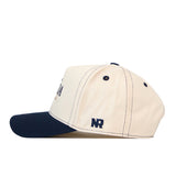 Introducing the No Rivals Nevada Wolf Pack Vintage Hat—a cream-colored baseball cap featuring a navy blue brim, navy stitching, and two navy eyelets. The initials “NR” are embroidered in navy thread near the back on the side. With a snap back adjustment for a comfortable and customizable fit, this hat is shown from a side view.