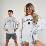 A woman poses confidently in a gray No Rivals Vanderbilt Mock Neck sweatshirt and matching shorts, while a man in an oversized fit Michigan State sweatshirt and gray shorts stands in the background against a plain white backdrop.