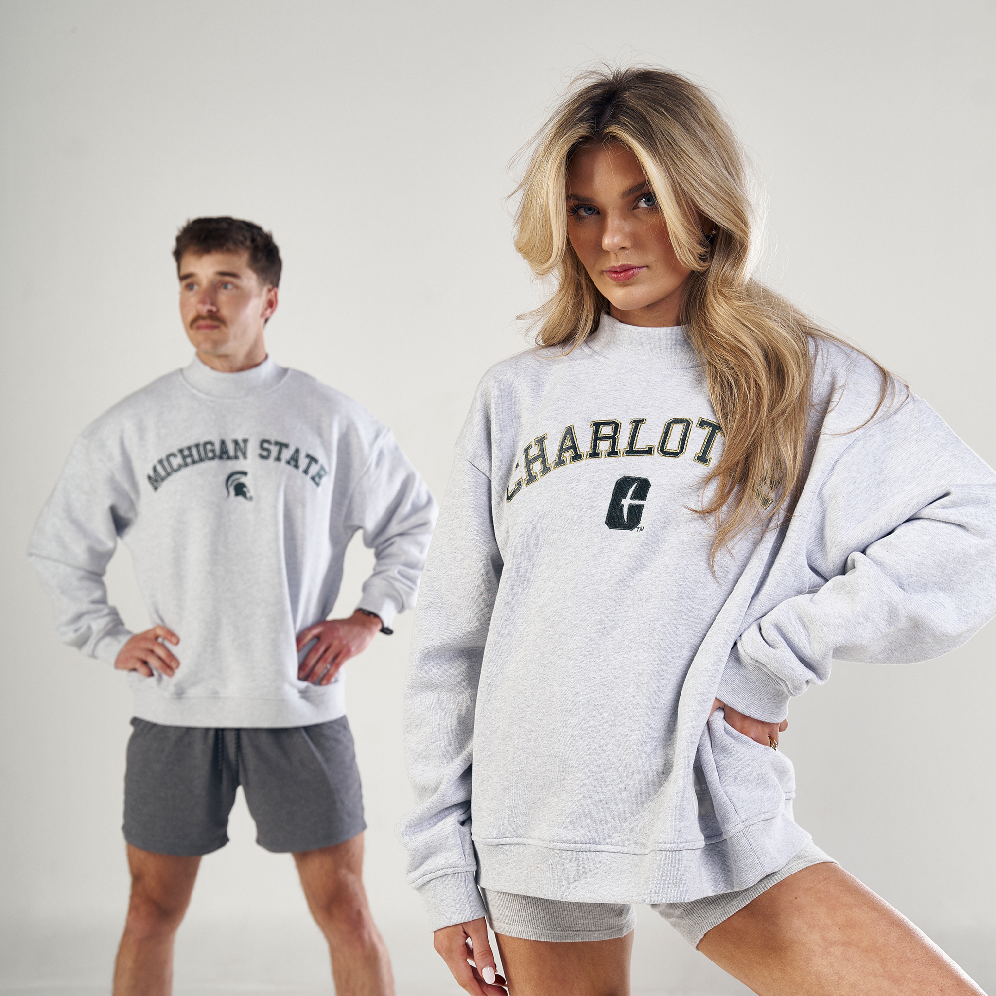 A woman poses confidently in a gray No Rivals Vanderbilt Mock Neck sweatshirt and matching shorts, while a man in an oversized fit Michigan State sweatshirt and gray shorts stands in the background against a plain white backdrop.