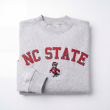 The No Rivals NC State Mock Neck is a folded gray heavyweight cotton sweatshirt featuring bold red NC STATE text, a central wolf mascot, and "NO RIVALS" in white on the cuff.