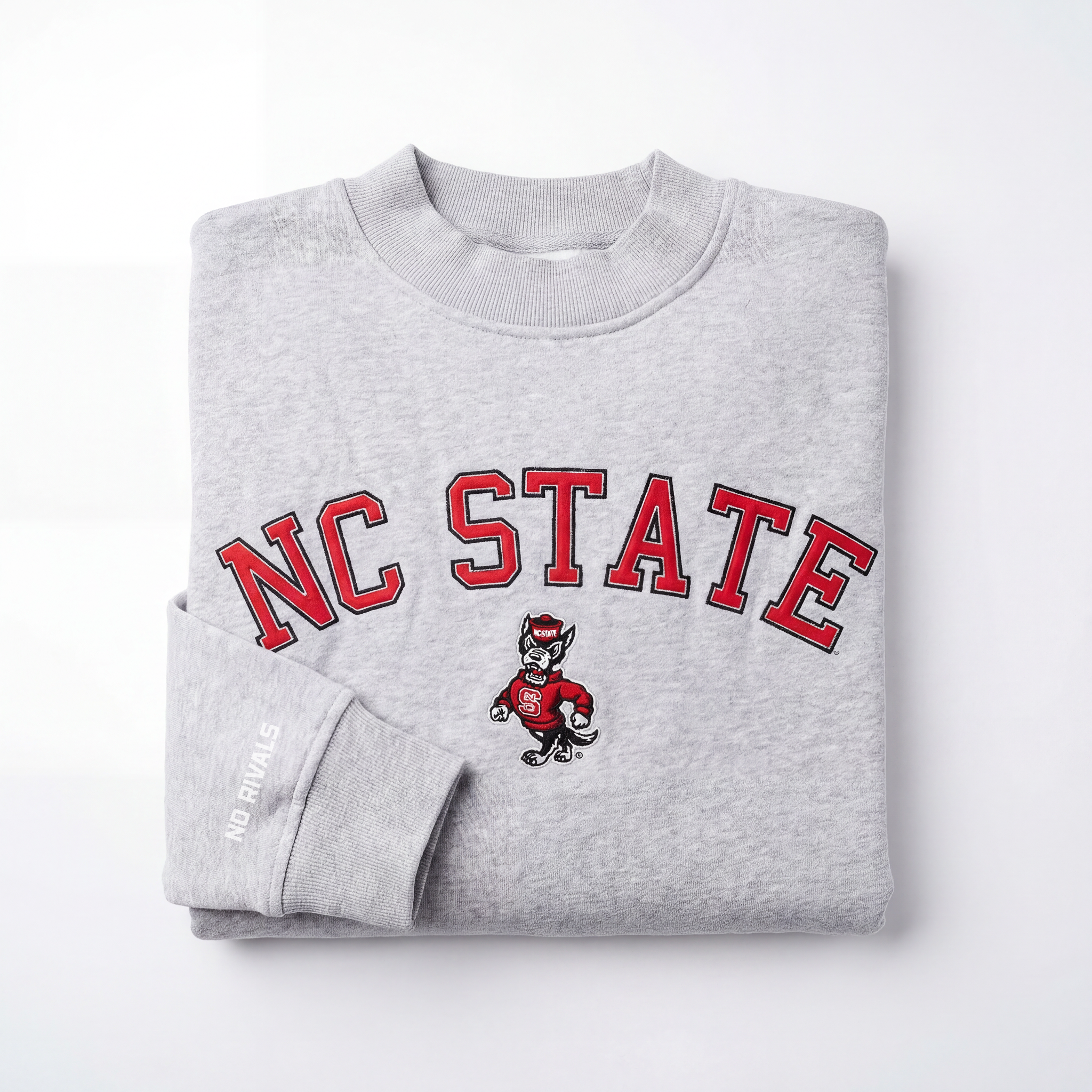 The No Rivals NC State Mock Neck is a folded gray heavyweight cotton sweatshirt featuring bold red NC STATE text, a central wolf mascot, and "NO RIVALS" in white on the cuff.