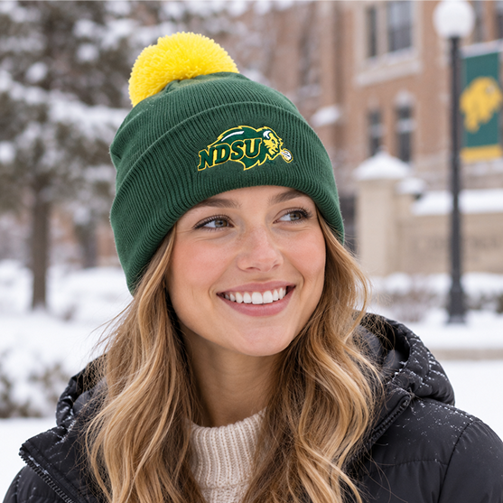 A smiling woman stands outside in the snow, wearing a No Rivals North Dakota State Bison Logo Beanie in green and yellow with an embroidered logo, along with a black jacket. A brick building and NDSU banners are visible in the background.