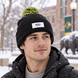 A young man stands outside in the snow, wearing a black No Rivals North Dakota State Bison Patch Beanie and jacket. He showcases the beanie’s embroidered logo, highlighting the officially licensed gear, with campus buildings and a bison statue visible in the background.