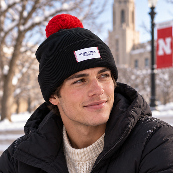A person wearing a No Rivals Nebraska Cornhuskers Patch Beanie in black with an embroidered logo and a black jacket sits outdoors in the snow, with a blurred building and red Nebraska banner in the background.