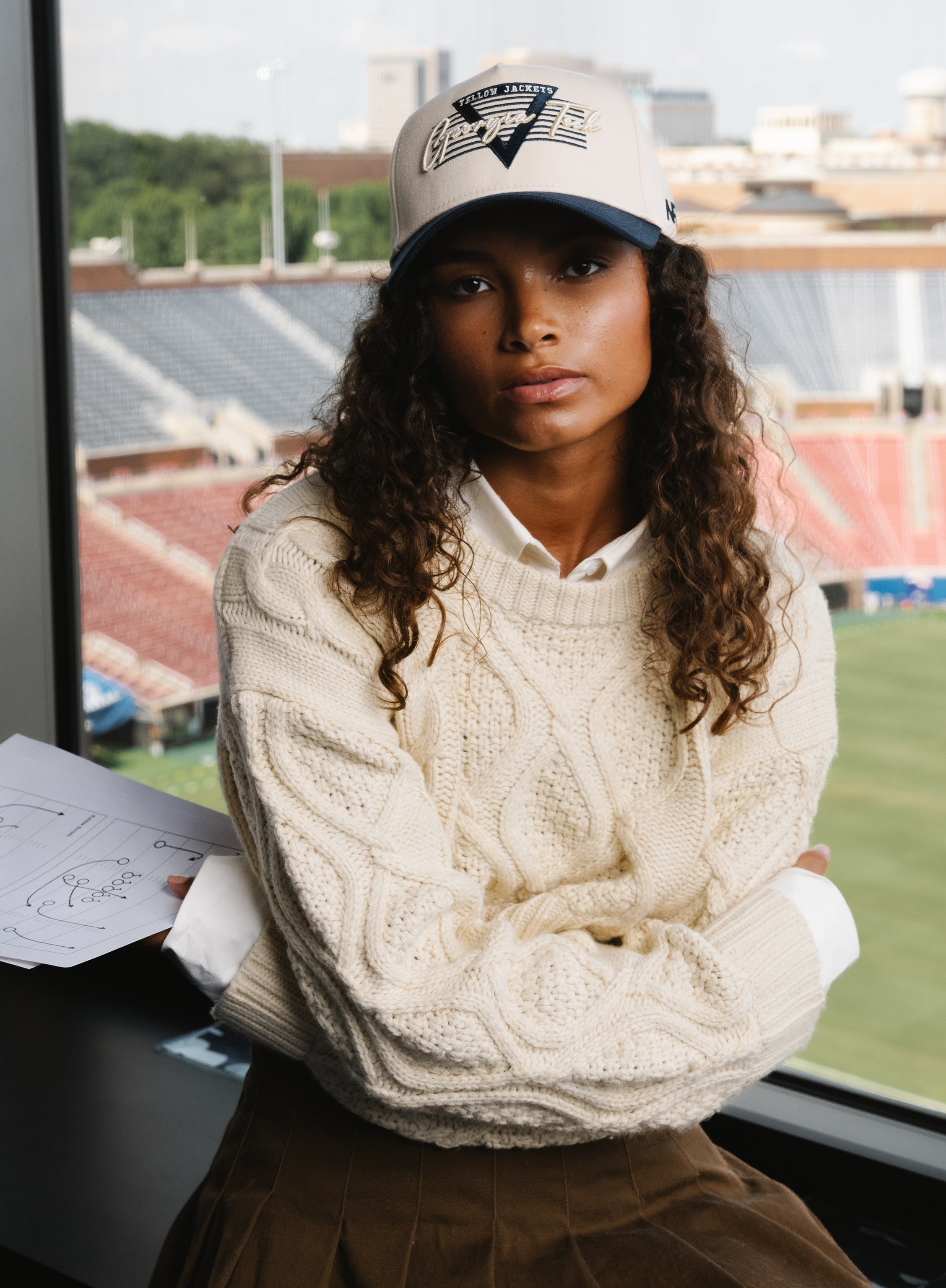 Person wearing a cream cable knit sweater and cap with stadium in the background