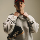 A man with a mustache holds a shoe while wearing the Takedown Mockneck - Ash Grey - PRE-SALE, styled in an oversized fit.