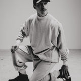 Wearing the Takedown Mockneck in Ash Grey (PRE-SALE), a model kneels on one knee in a minimal studio. The black and white photo highlights the casual, oversized fit and stylish, understated mood of the heavyweight cotton sweatshirt.