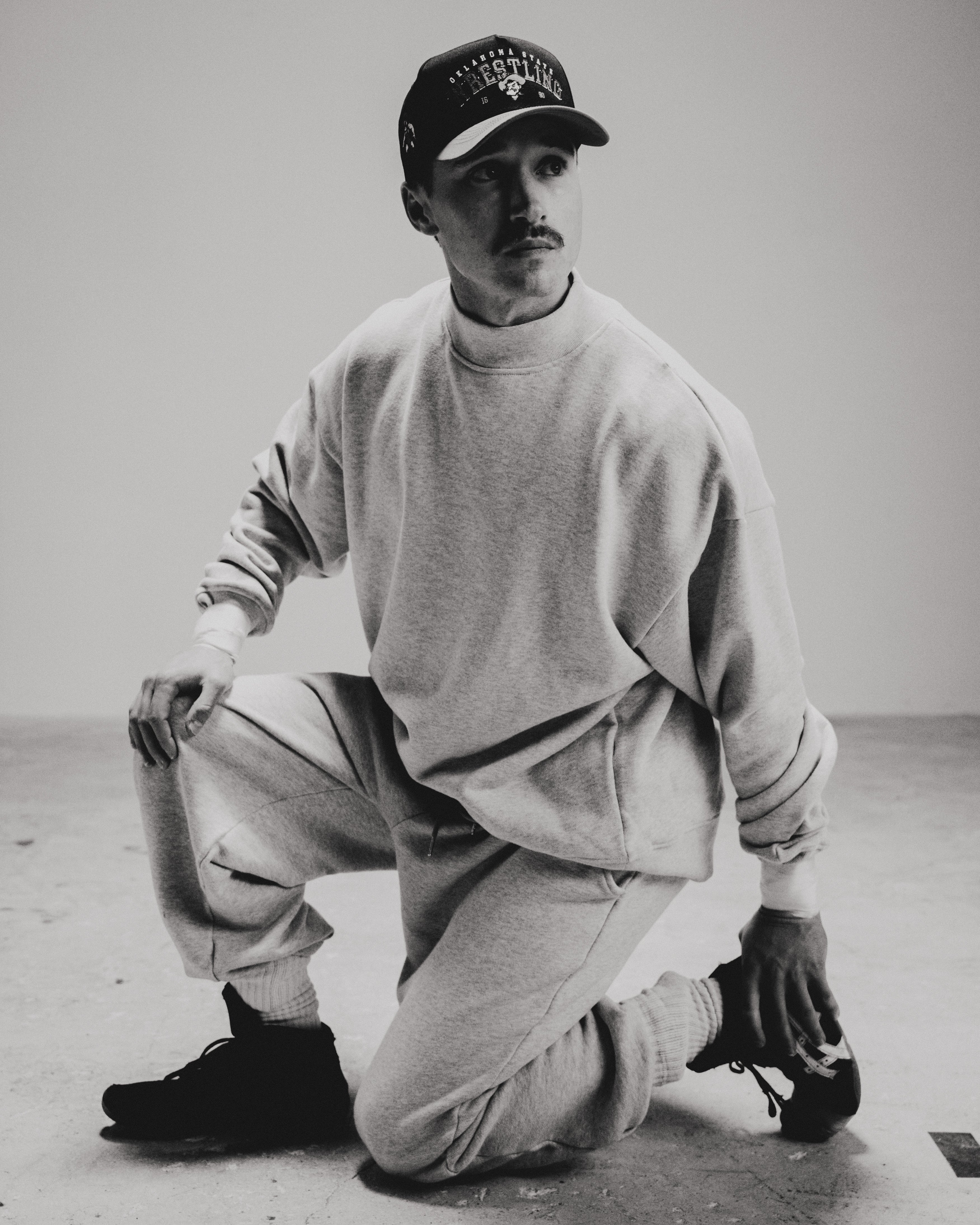 Wearing the Takedown Mockneck in Ash Grey (PRE-SALE), a model kneels on one knee in a minimal studio. The black and white photo highlights the casual, oversized fit and stylish, understated mood of the heavyweight cotton sweatshirt.
