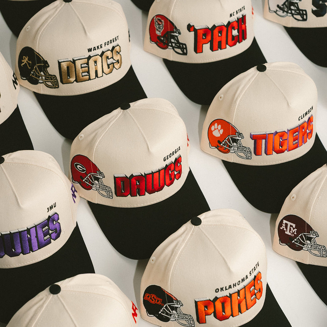 A collection of cream and black baseball caps, each featuring embroidered logos and mascots of various college football teams, such as the Dawgs, Tigers, Deacs, Pokes, and Vols, arranged in neat rows.