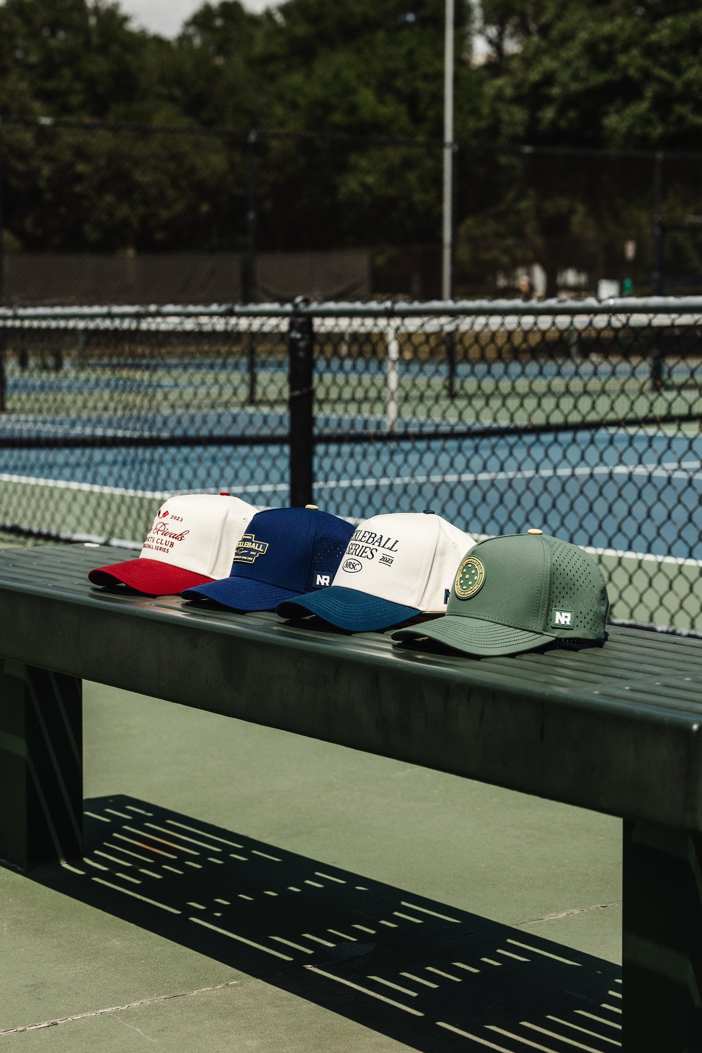 Four No Rivals hats from the Pickleball Drop sit on a green bench in front of a blue tennis court. The hats include two athletic mesh styles and two cotton builds, each with unique pickleball-themed designs.