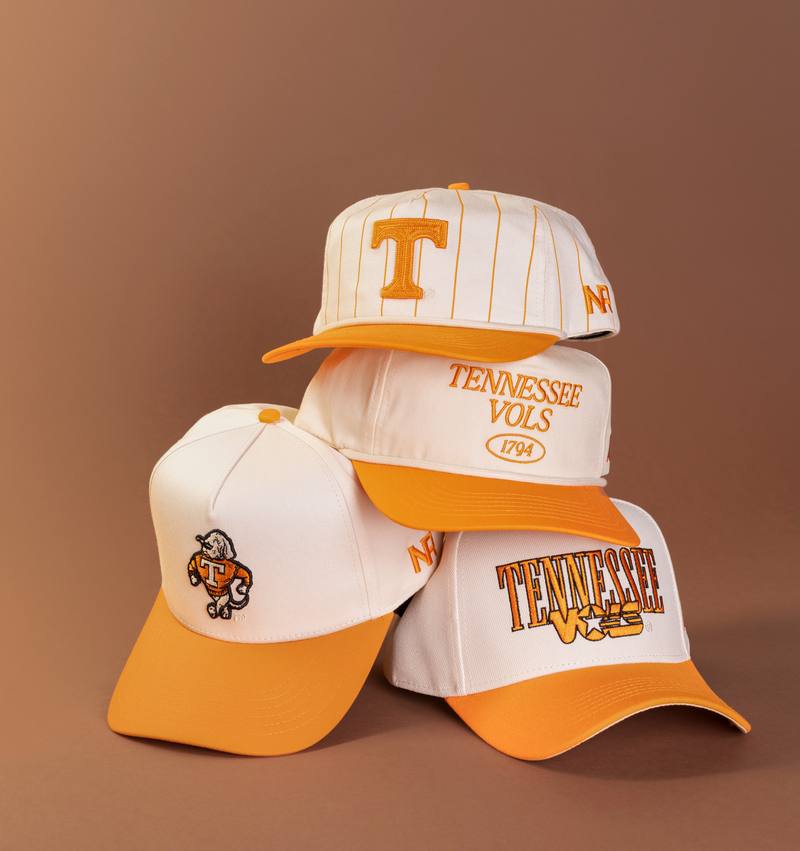 Four white and orange Tennessee Vols baseball caps are stacked on a brown background, each displaying different logos and text related to the University of Tennessee.