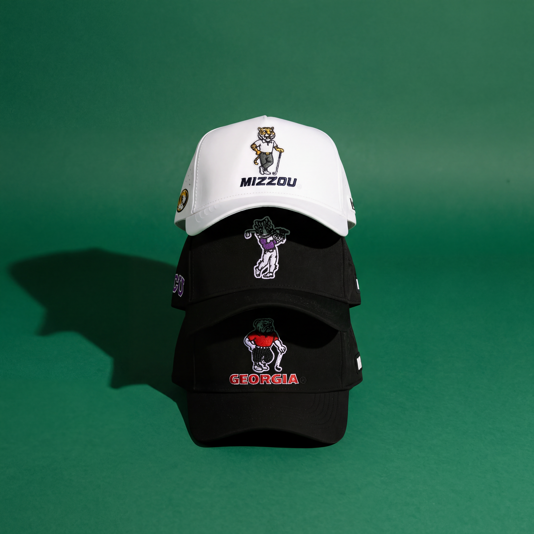 Three baseball caps stacked vertically on a green background. The top hat is white with MIZZOU and a tiger mascot, while the two hats below are black with embroidered mascots and GEORGIA text in red on the bottom cap.