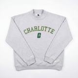 The No Rivals UNCC Mock Neck is a light gray, heavyweight cotton crewneck with "CHARLOTTE" in bold green and yellow letters across the chest, a green logo underneath, and an oversized fit, shown on a white background.