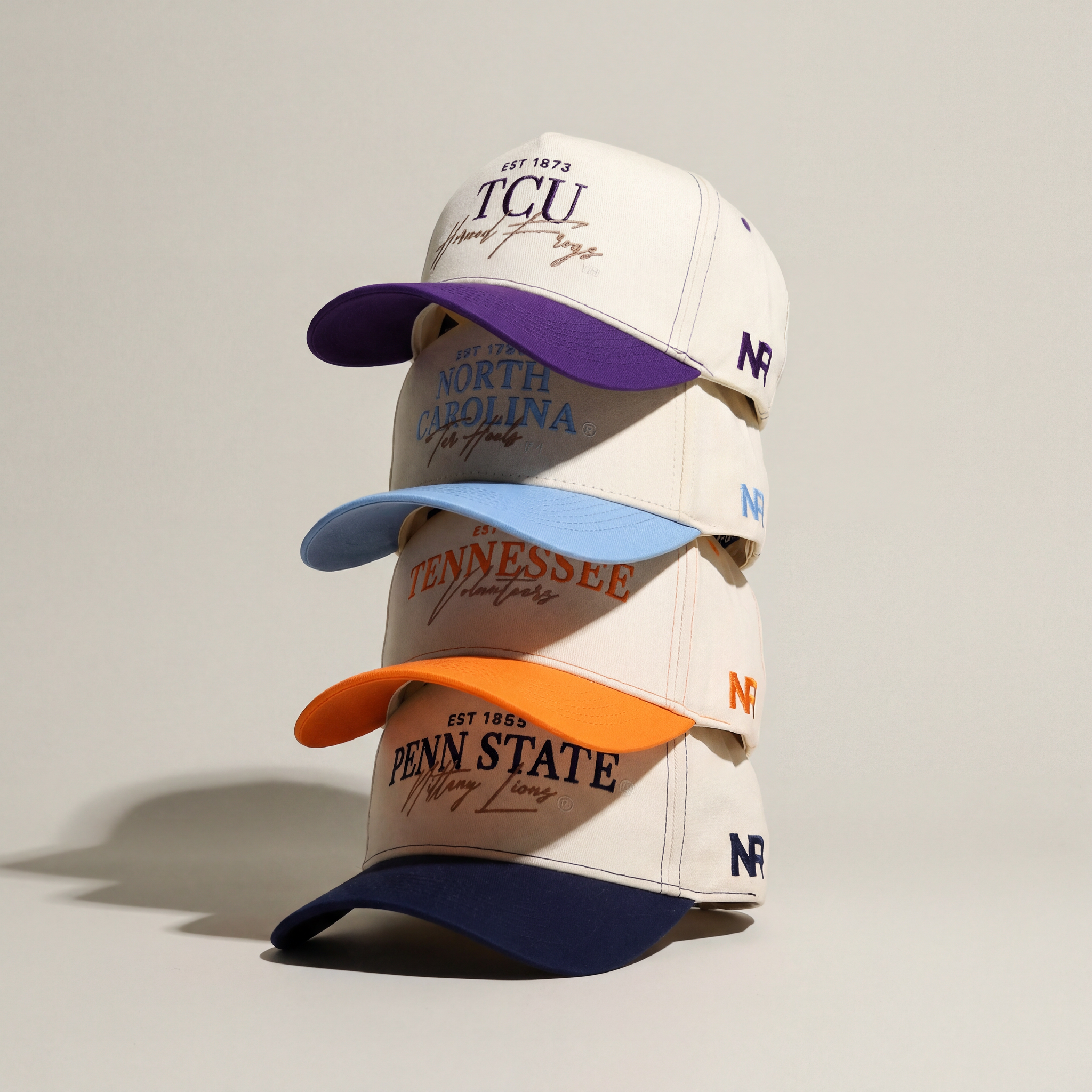 Four white baseball caps are stacked vertically. Each features a different university name: TCU (purple brim), North Carolina (light blue brim), Tennessee (orange brim), and Penn State (navy brim), all on a plain background.