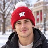 A young man smiles outdoors on a snowy day, wearing a No Rivals Wisconsin Badger Logo Beanie—a red beanie featuring an embroidered logo—paired with a black jacket, with a university building and trees in the background.